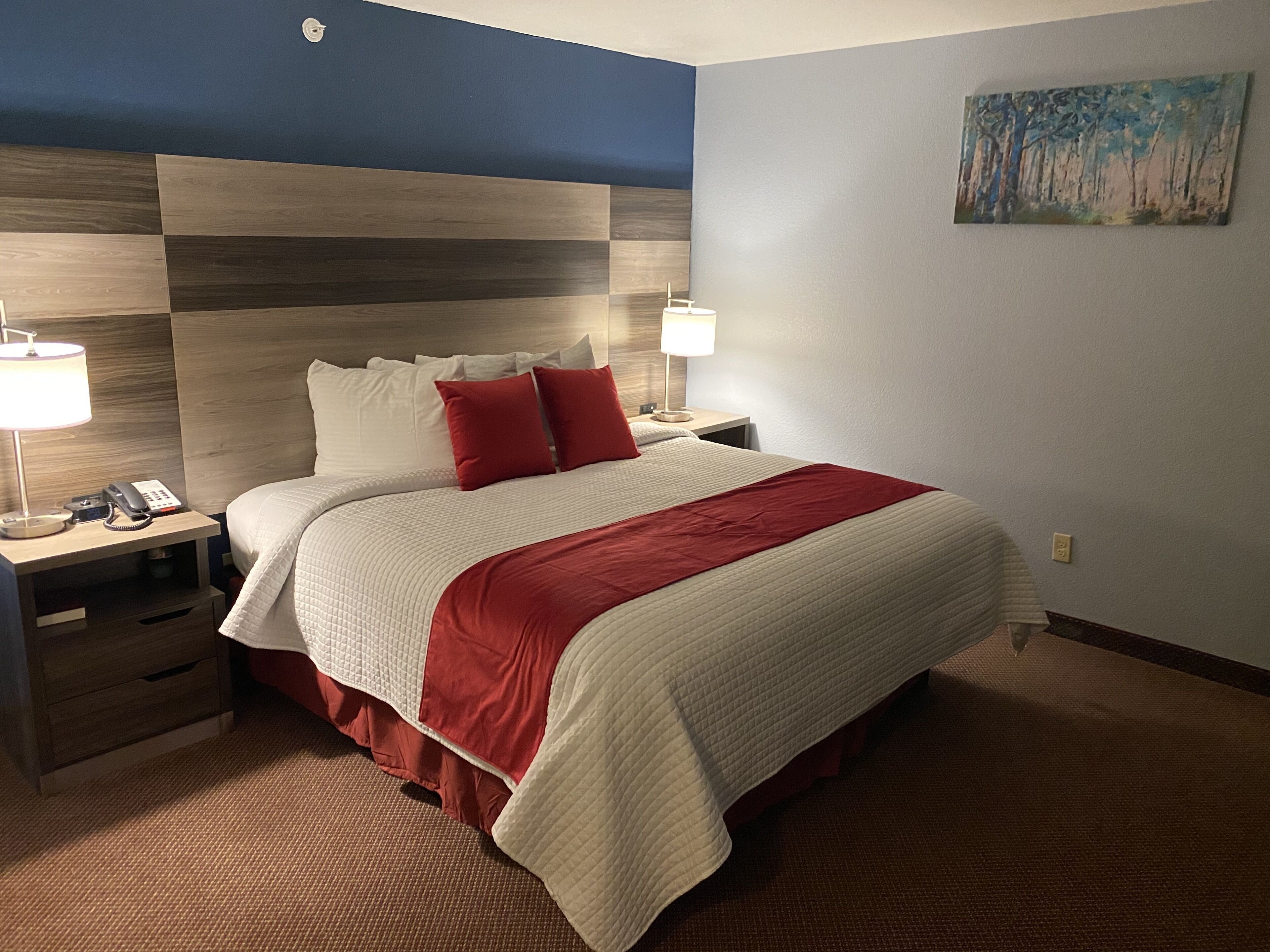 Una habitación de hotel con una cama grande, dos mesitas de noche, un revistero y una colcha roja y blanca, además de un cuadro en la pared.