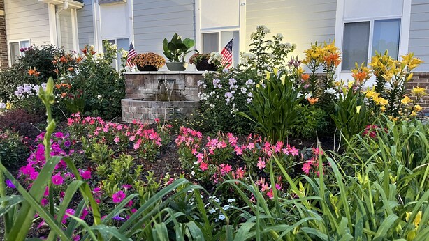 A garden with various flowers and plants in front of a building with a stone fountain.