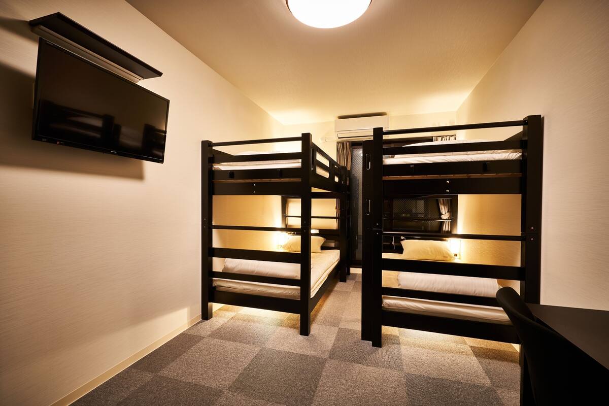 A dormitory room with bunk beds, a flat-screen TV mounted on the wall, and a ceiling light.