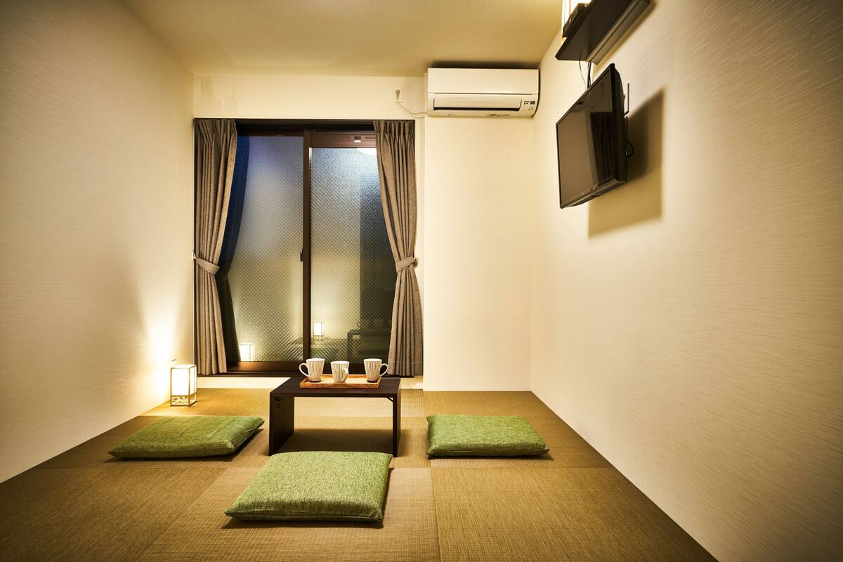 A room with tatami flooring, a low table with cups, a TV mounted on the wall, and a window with curtains.