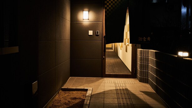 Un couloir étroit et faiblement éclairé, avec un motif à carreaux sur le mur, un petit tapis sur le sol et une porte surmontée d’une lumière.