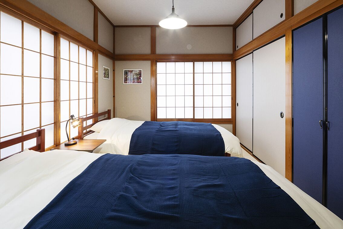 Et traditionelt japansk værelse med tatamimåtter, to senge med blåt sengetøj, en træsenggavl, et natbord med en lampe og skydedøre.