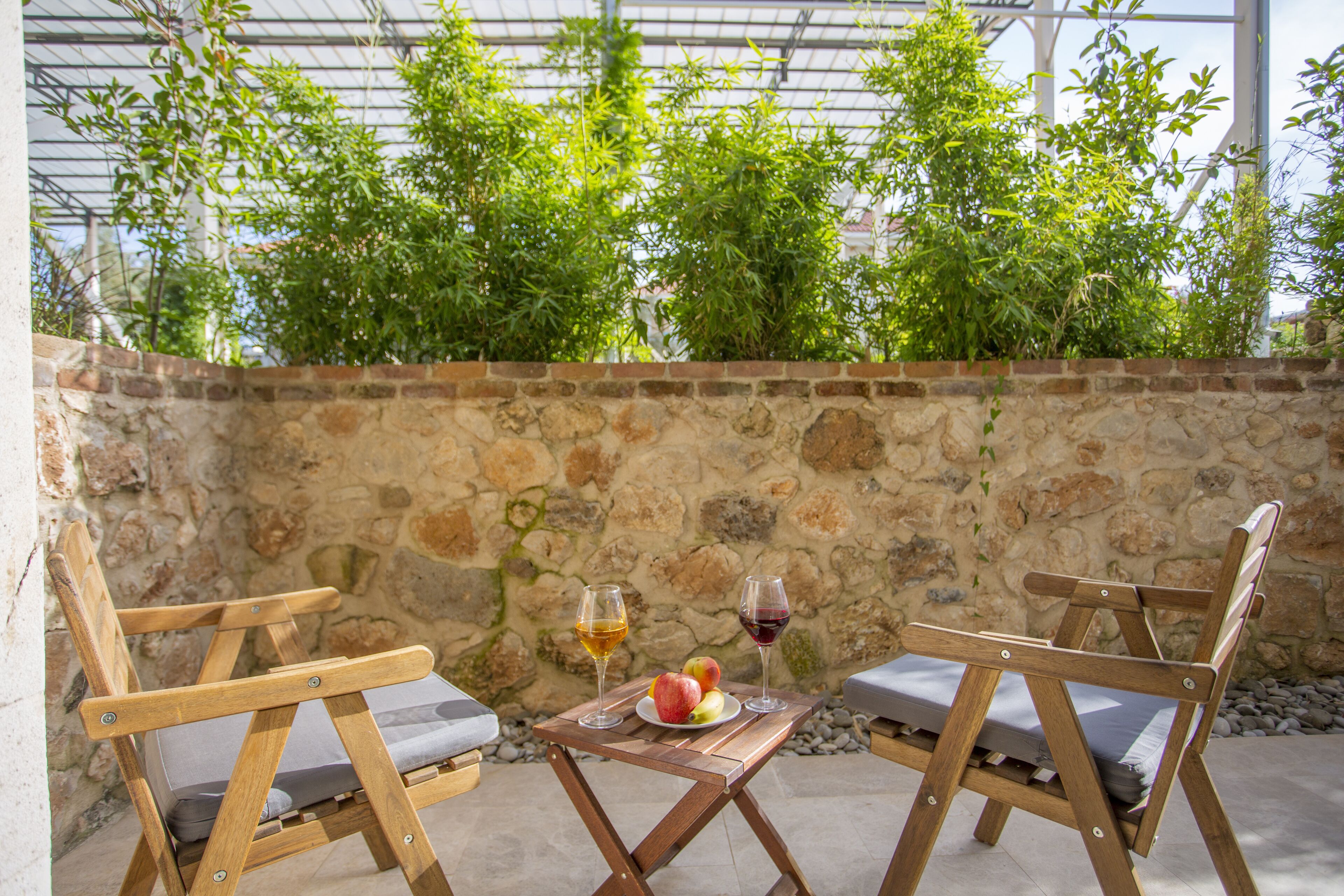 Deux chaises en bois avec des coussins, une table sur laquelle se trouve une assiette de fruits et deux verres de vin, et un mur de pierre avec de la verdure en arrière-plan.