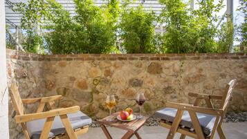 Deux chaises en bois avec des coussins, une table sur laquelle se trouve une assiette de fruits et deux verres de vin, et un mur de pierre avec de la verdure en arrière-plan.