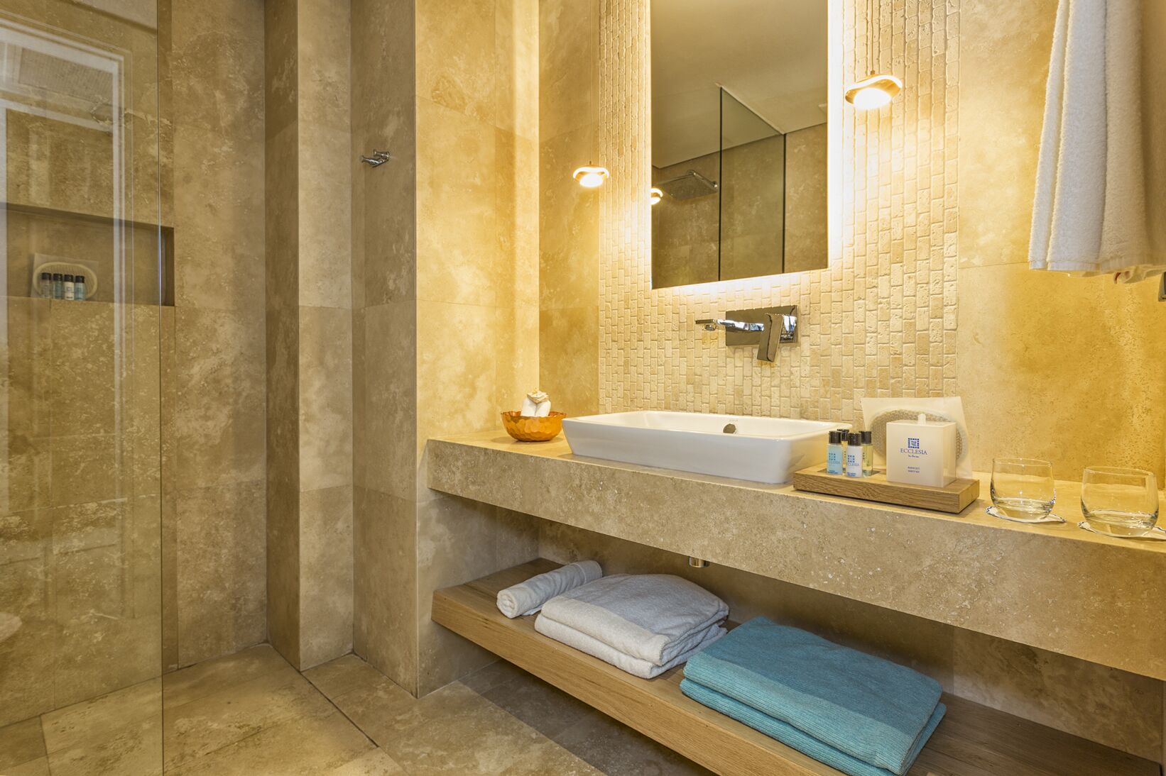 A modern bathroom with a glass-enclosed shower, a large sink, and a mirror above it. There are towels and toiletries on the counter.
