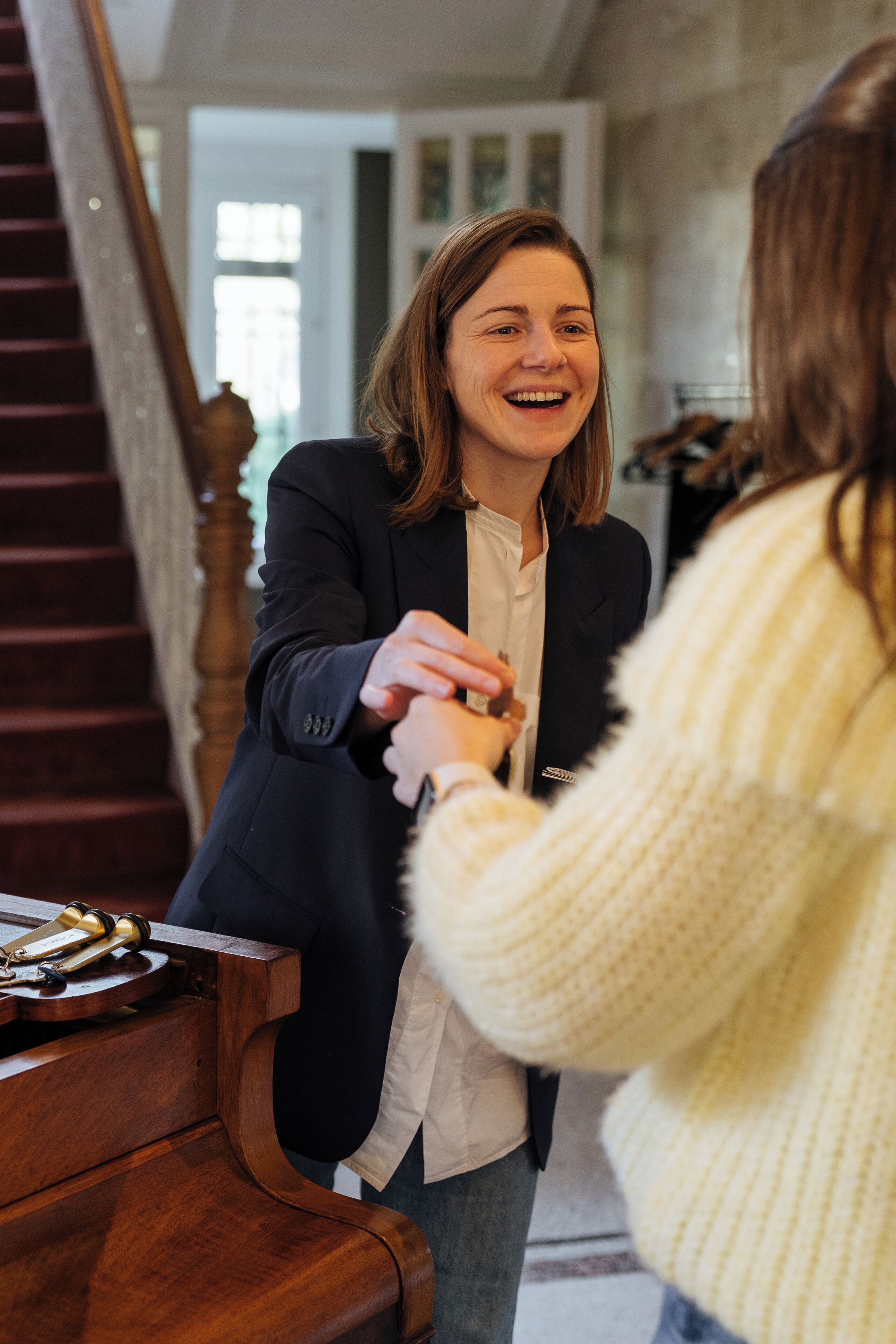 Une personne vêtue d’un blazer sombre et d’une chemise blanche se tient près d’un piano en bois, tenant la main d’une autre personne qui porte un pull de couleur claire.