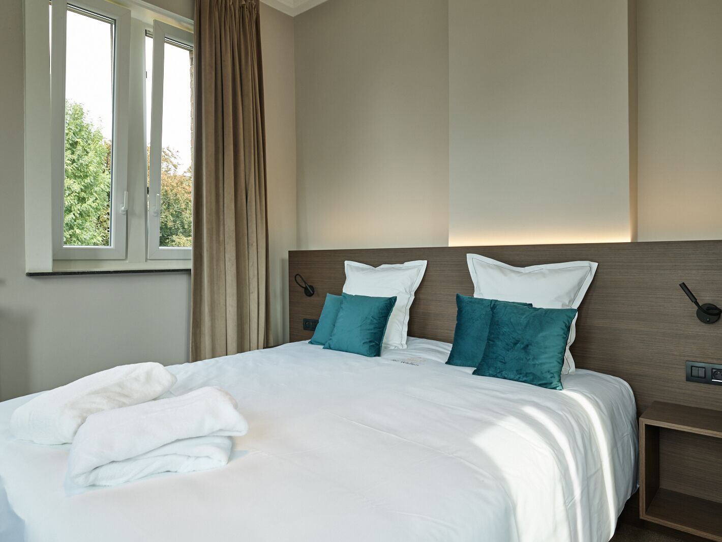 Un lit bien fait, avec du linge de lit blanc et des coussins turquoise, une tête de lit en bois, une table de chevet avec une lampe et une fenêtre avec des rideaux.