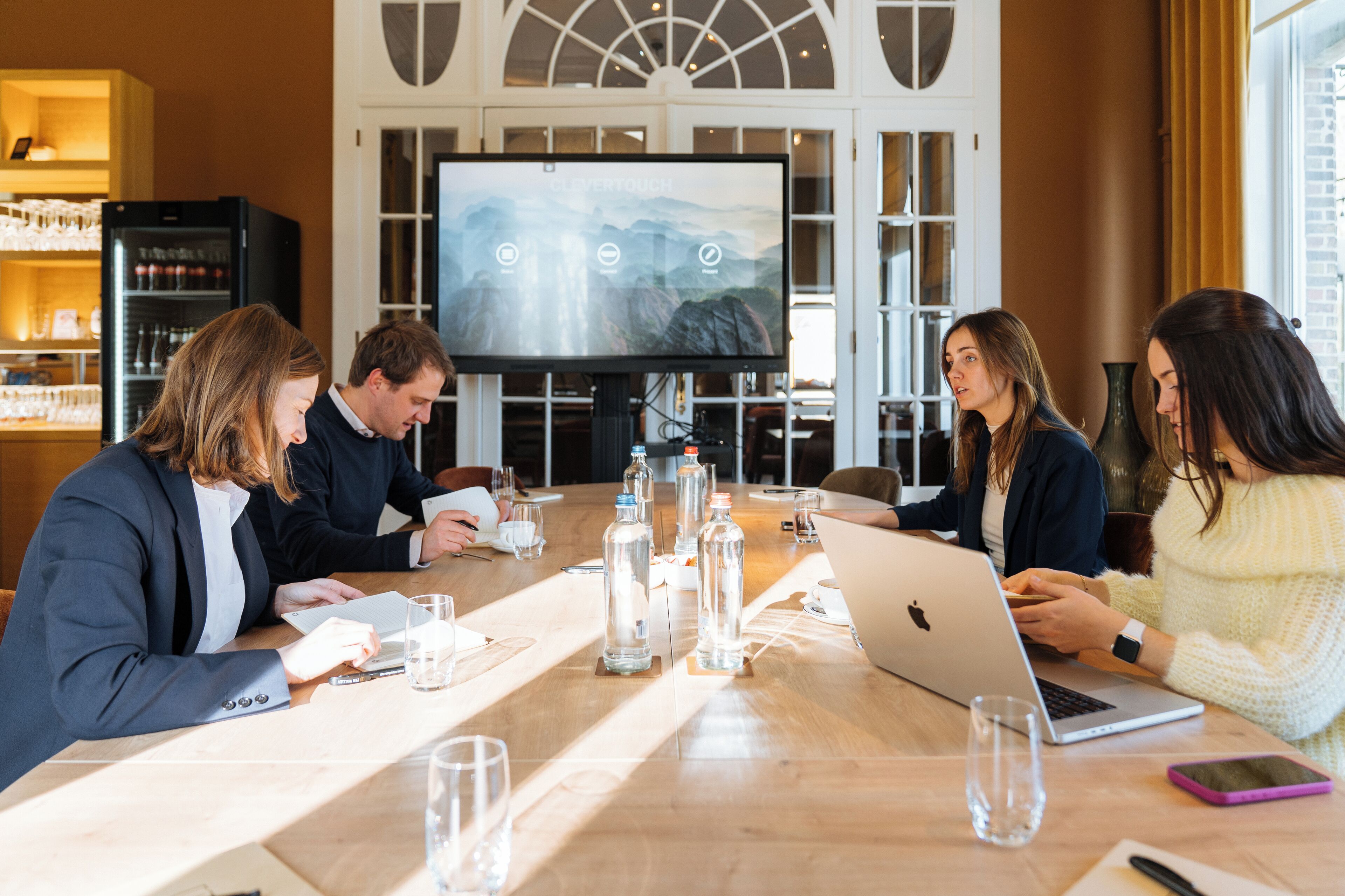 A conference room with a large screen, a wooden table, and several people engaged in a meeting.