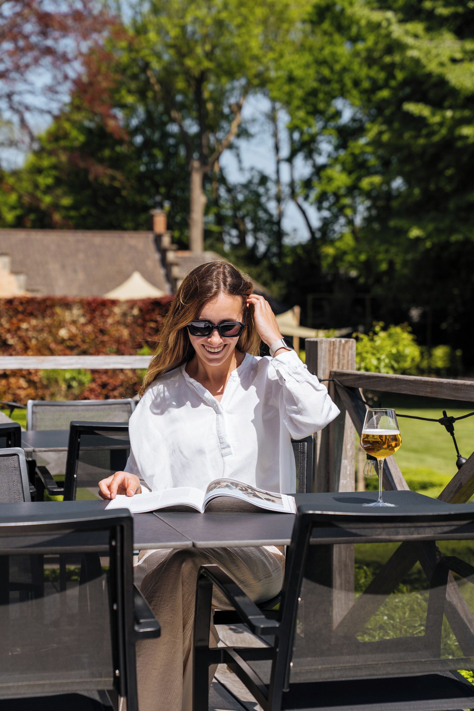 Una persona sentada en una mesa al aire libre, con un libro, una copa de vino y vistas a árboles y un edificio al fondo.