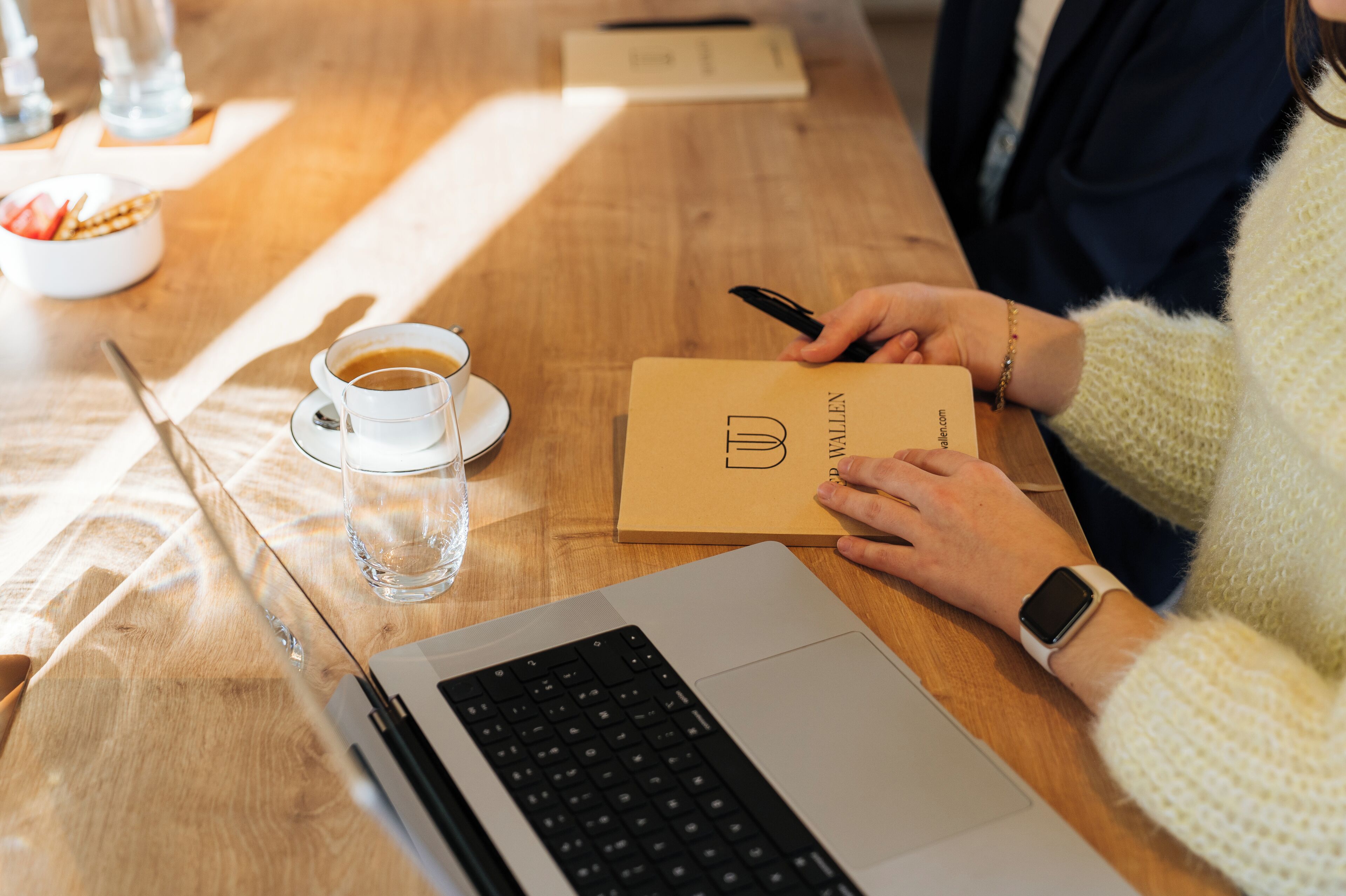 Una mesa de madera con una computadora portátil, una taza de café, un vaso de agua, un cuenco de fruta y una persona sosteniendo una tarjeta.