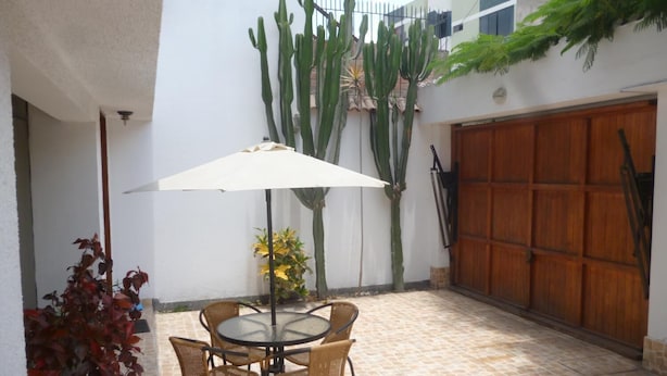 Uma área de pátio com mesa redonda e cadeiras, guarda-sol branco e cactos em frente a uma parede branca.