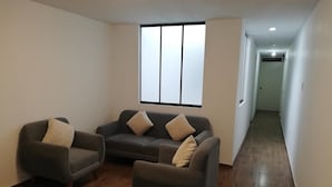 Quarto com sofá cinzento e duas poltronas, uma janela grande com persianas, soalho de madeira e um corredor que dá para outro espaço.
