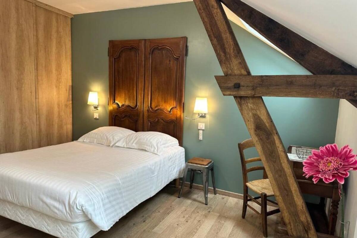 A bedroom with a bed, wooden headboard, two wall-mounted lamps, a small table, a chair, and a flower vase.