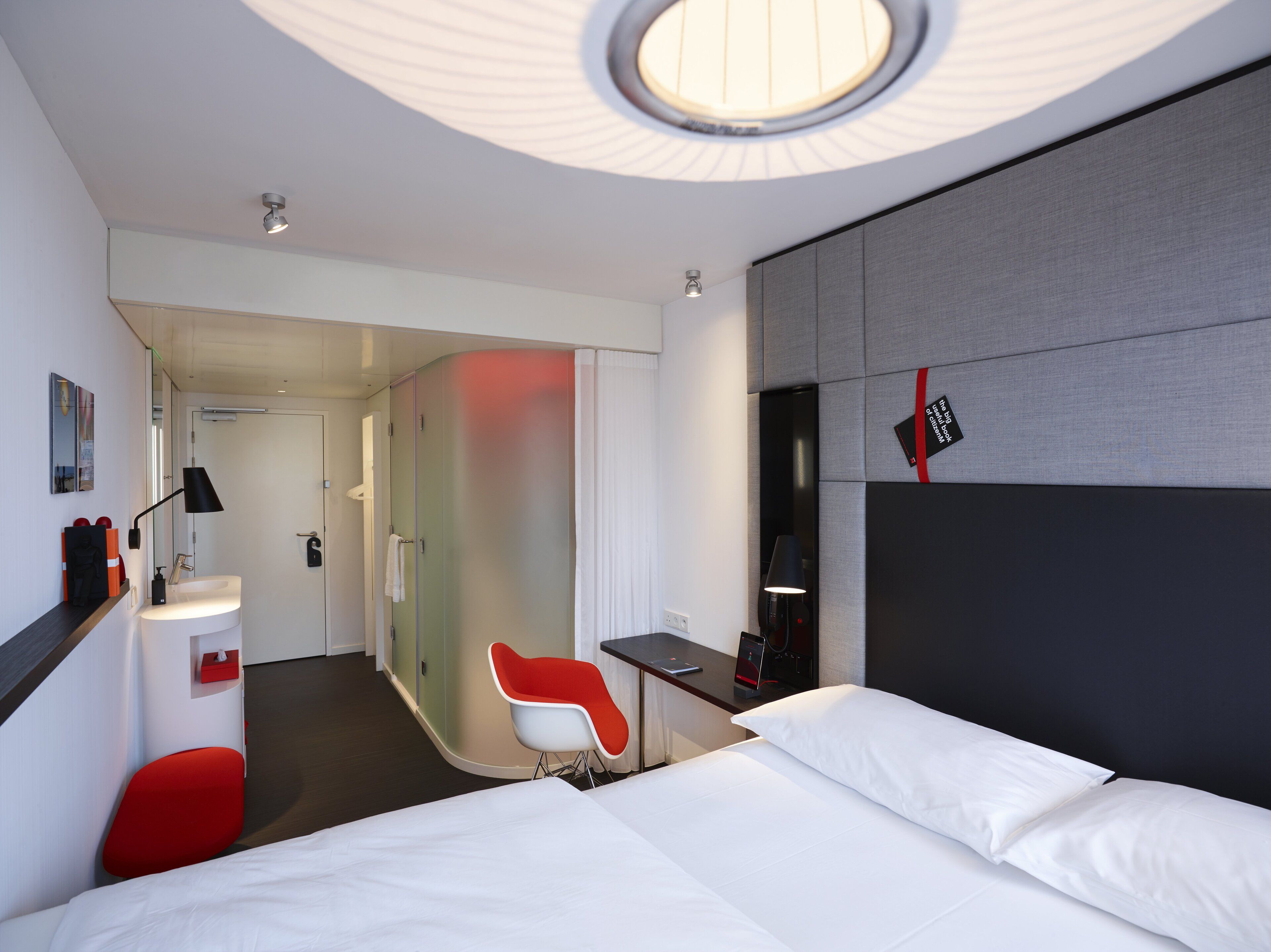 A modern hotel room with a large bed, a red chair, a desk, and a bathroom visible through a glass partition.
