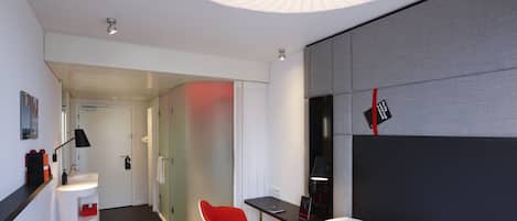 A modern hotel room with a large bed, a red chair, a desk, and a bathroom visible through a glass partition.