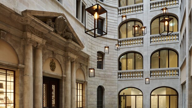 Un amplio patio con varias mesas y sillas, rodeado por un edificio de estilo clásico con ventanas de arco y fachada de piedra.