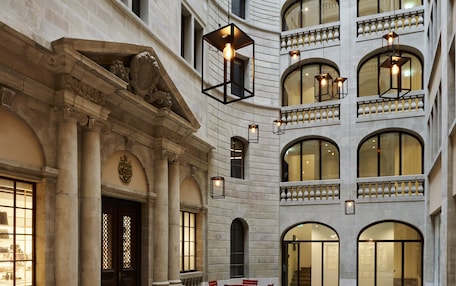 A spacious courtyard with multiple tables and chairs, surrounded by a classical building with arched windows and a stone facade.