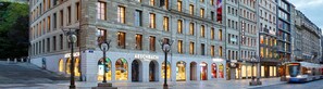 A multi-story building with a row of storefronts, including a visible 'AESC BACH' store. - citizenM Geneva (Geneva)