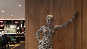 Una escultura de una mujer con vestido de pequeñas cuentas, ubicada en un vestíbulo con paneles de madera y techo curvo.