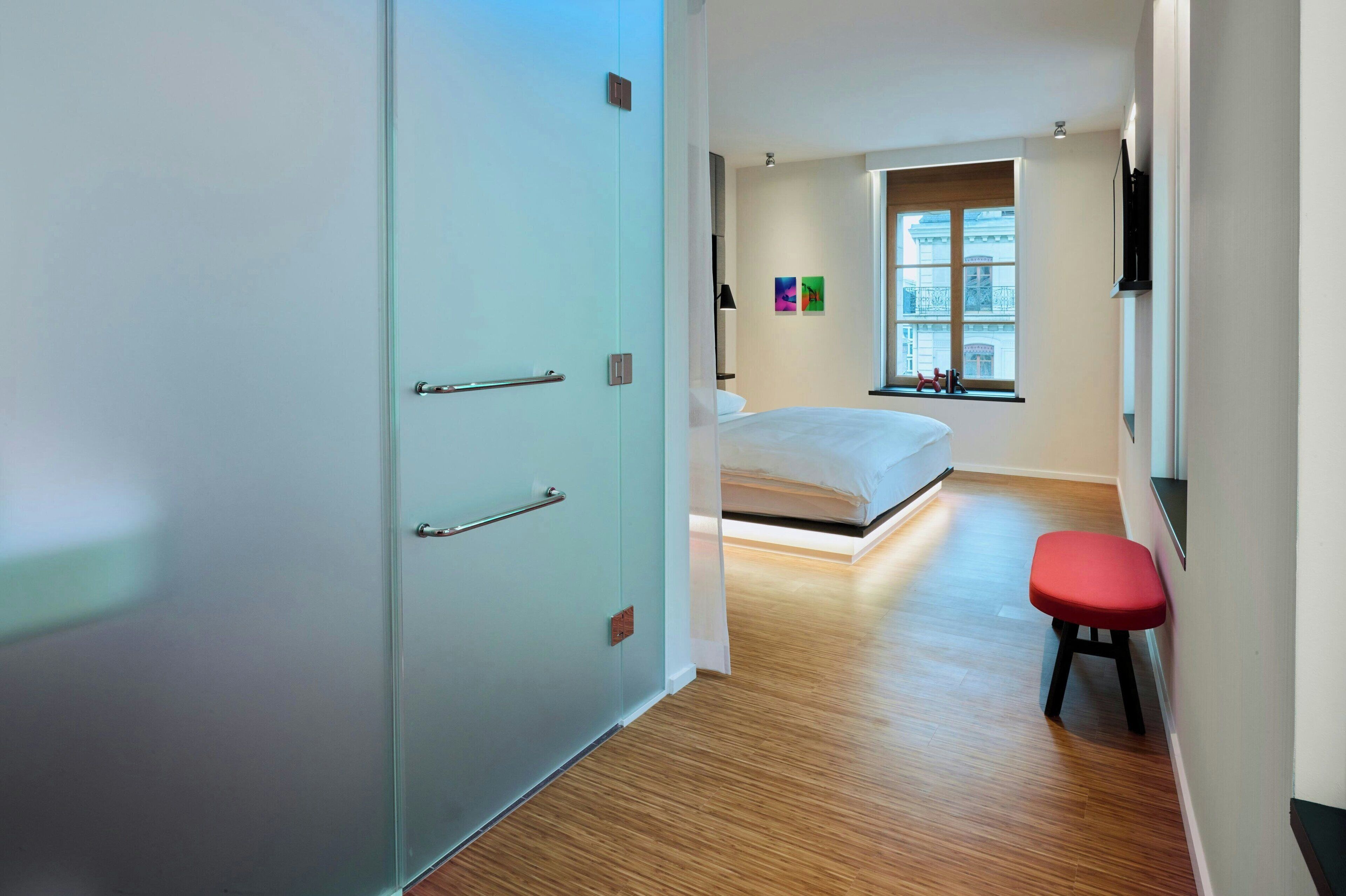 A modern hotel room with a bed, a red stool, a window with a view, and a door with a handle.