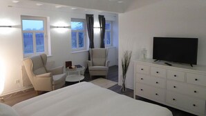 Habitación de hotel moderna con una cama grande, dos sillones, una mesita, un televisor en una cómoda y ventanas amplias con cortinas.