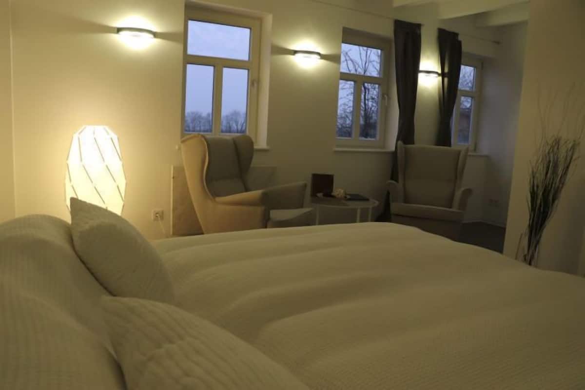 A hotel room with a large bed, two armchairs, a small table, and a lamp. There are windows with curtains and a view of trees outside.