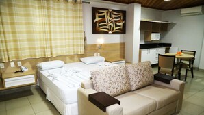 A hotel room with a bed, a sofa, a desk, a chair, a small table, and a kitchen area with a microwave. - Swamy Hotel (Cruzeiro do Sul)