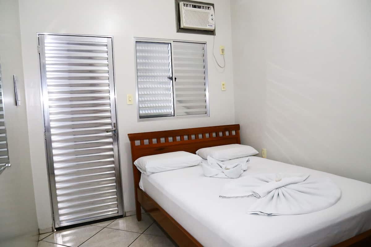 Une chambre d’hôtel avec un lit, une tête de lit en bois, deux fenêtres équipées de stores, un radiateur et une porte.