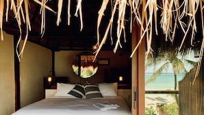 A bedroom with a wooden bed, white bedding, a round mirror, and a view of the beach. - Libelula Tulum Beachfront Hotel (Tulum)