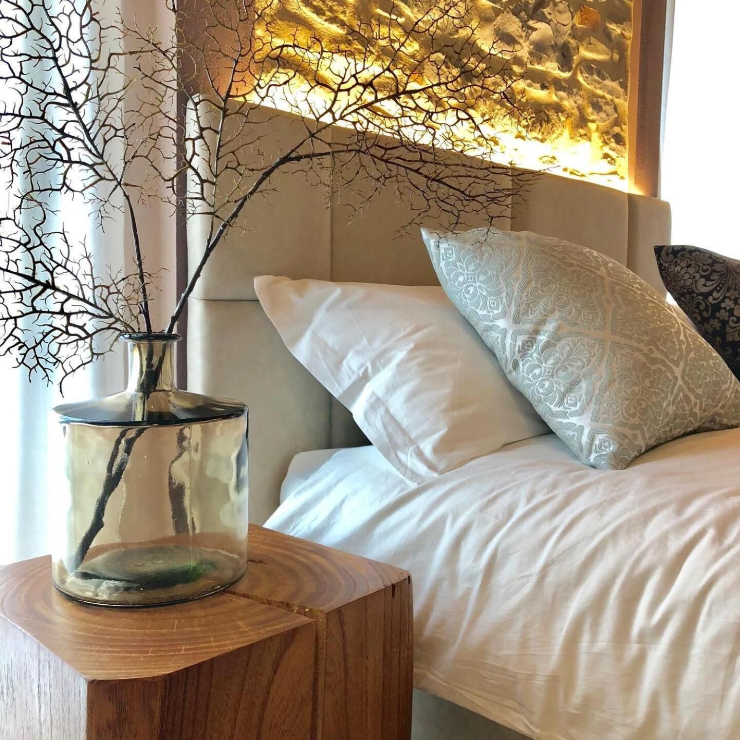 Un dormitorio con cama, ropa de cama blanca, un jarrón decorativo con ramas y una almohada estampada.