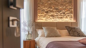 Ein Schlafzimmer mit einer Wand aus Stein, einem Bett mit gemusterten Kissen und einer Vase mit Zweigen auf einem Nachttisch.
