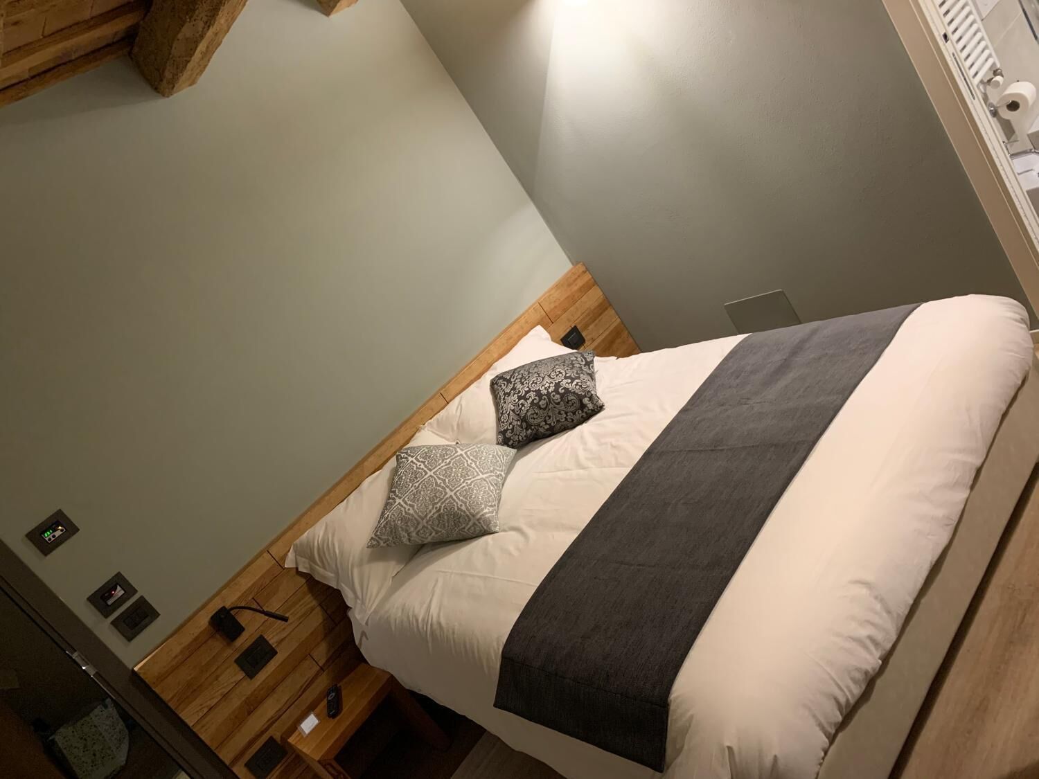 Una cama bien hecha con una manta gris y almohadas estampadas, un cabecero de madera y una mesita de noche con una lámpara.