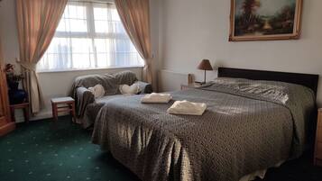 A bedroom with a large bed, a nightstand, a lamp, a window with curtains, a painting on the wall, and a small stool.