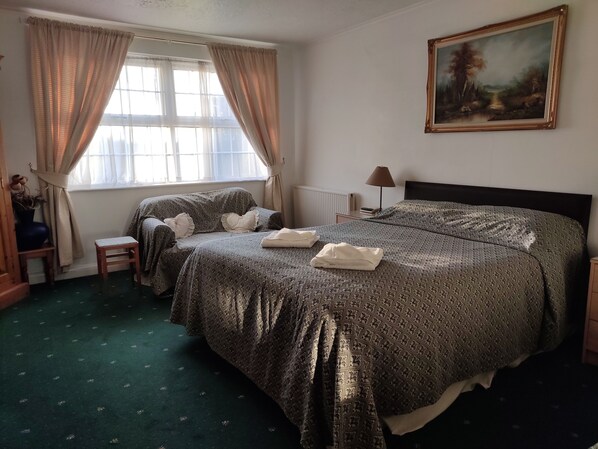 Un dormitorio con una cama grande, una mesita de noche, una lámpara, una ventana con cortinas, un cuadro en la pared y un taburete pequeño.