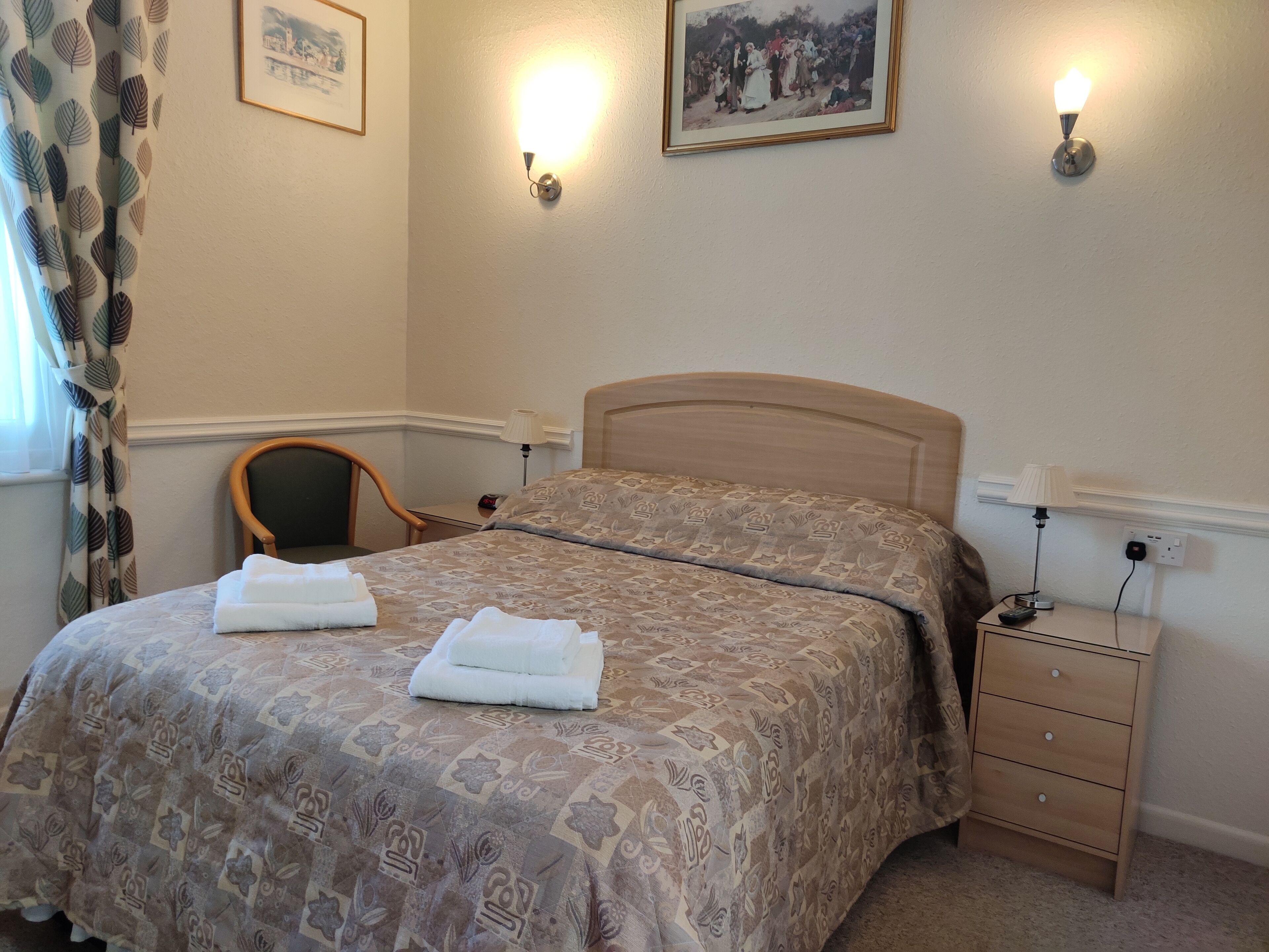 Cama bem arrumada com colcha estampada e duas toalhas dobradas. Há uma cadeira de madeira, uma mesa de cabeceira com abajur e um aparelho de iluminação fixado na parede.
