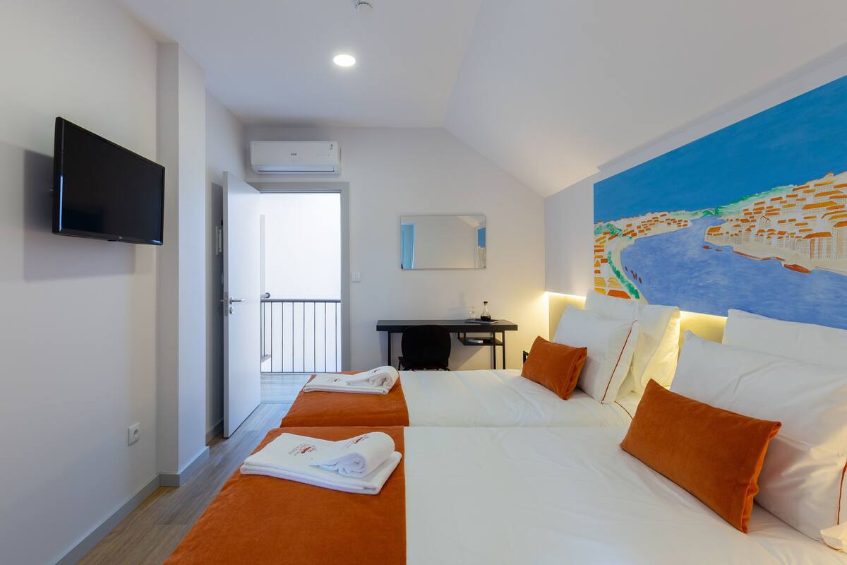 A hotel room with two beds, a desk, a TV, and a painting of a river scene.