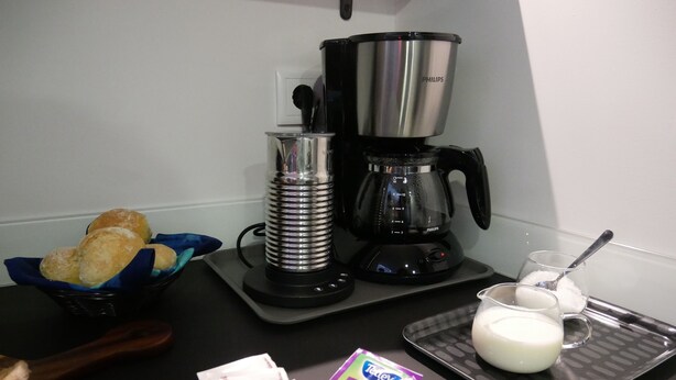 A kitchen counter with a coffee maker, a basket of bread, a plate with coffee packets, and a glass of milk.