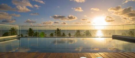 Piscina infinita en la azotea con terraza de madera, vista al atardecer y palmeras.