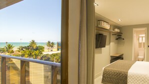 A hotel room with a balcony overlooking a beach, a bed with a quilted bedspread, a flat-screen TV mounted on the wall, and a small shelf with decorative items. - Infinity at the Sea by Welkom (João Pessoa)