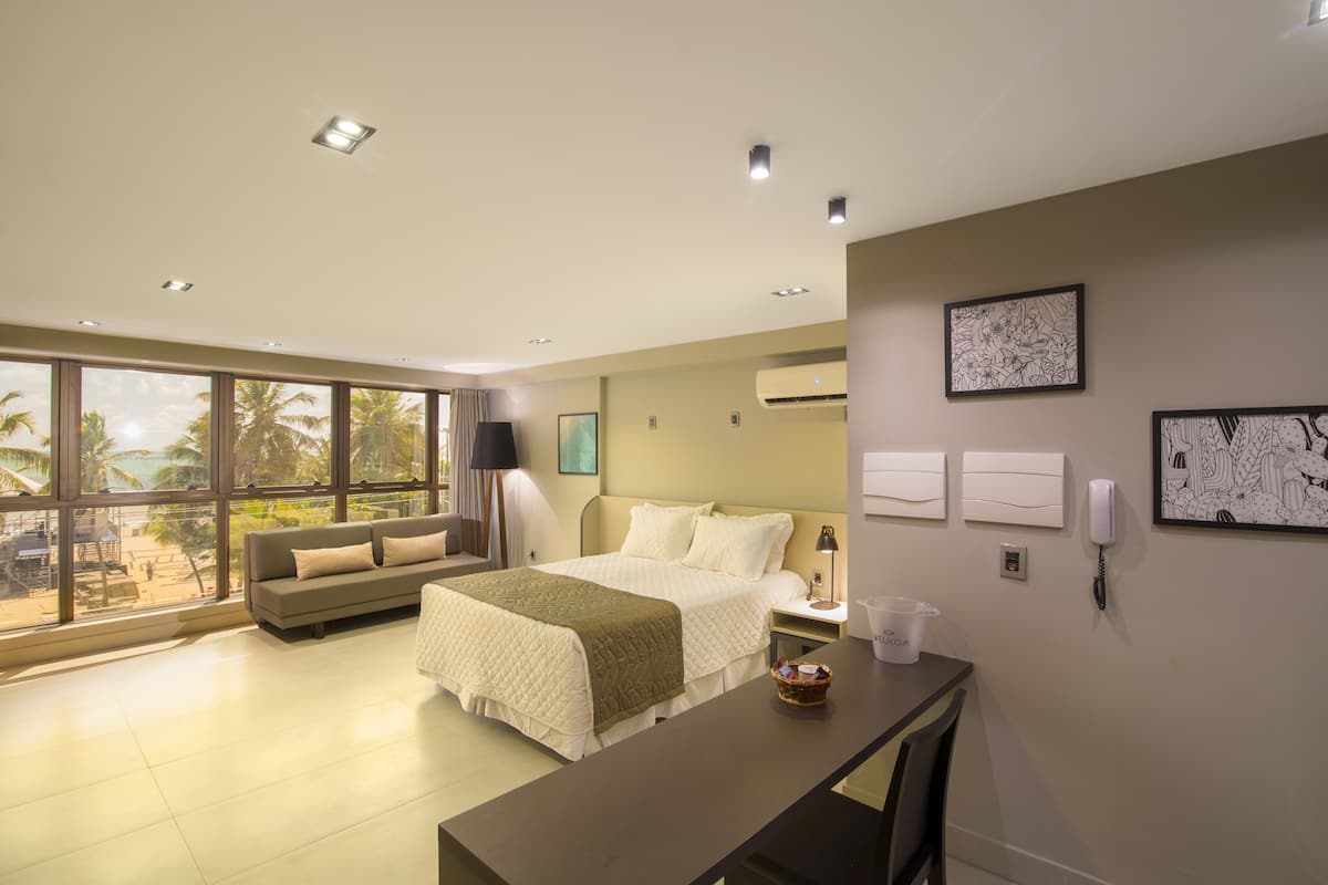 Ein modernes Hotelzimmer mit einem großen Bett, einer Sitzecke mit einer Couch, einem Schreibtisch und Blick auf Palmen durch große Fenster.