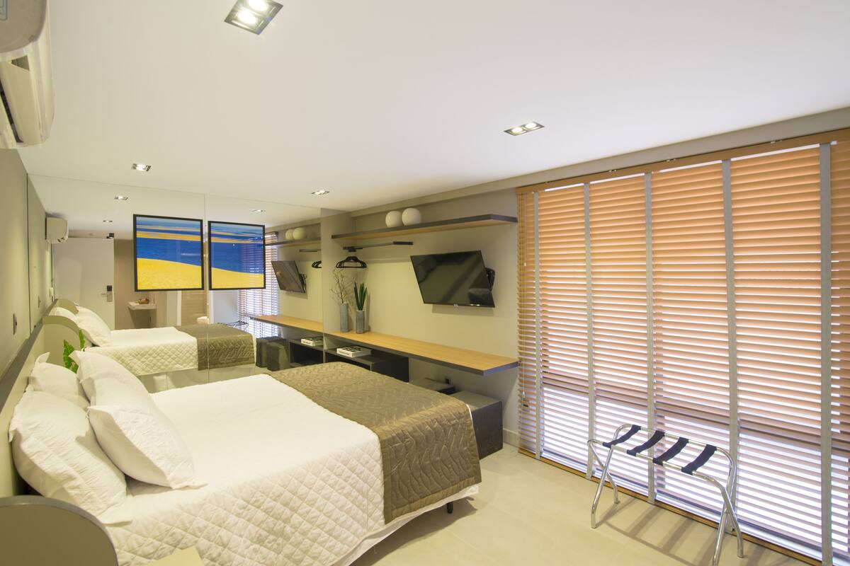 Ein modernes Hotelzimmer mit einem großen Bett, einem Schreibtisch mit Flachbildfernseher, einer Sitzecke mit einer Bank und einem großen Fenster mit Jalousien.