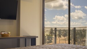 A bedroom with a large window overlooking a beach, a flat-screen TV mounted on the wall, and a bedside table with a basket. - Infinity at the Sea by Welkom (João Pessoa)