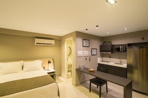 A compact hotel room with a bed, bedside table, and a kitchenette area including a refrigerator, microwave, and sink.