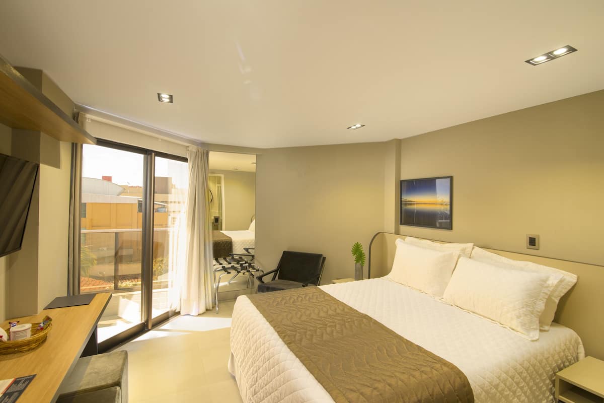 Ein Hotelzimmer mit einem großen Bett, einem Schreibtisch mit Stuhl, einem Fernseher und einem Balkon mit Blick auf Gebäude.
