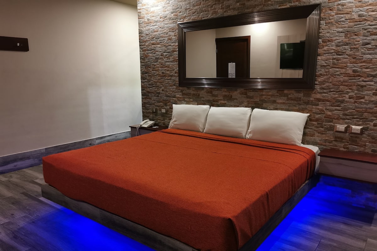 A modern bedroom with a stone wall, a large bed with an orange bedspread, a wooden nightstand, and a blue-lit floor.