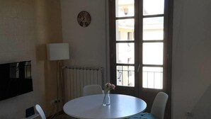 A room with a round table, two chairs, a lamp, a clock, and a window with a view of buildings. - Two-room apartment overlooking the Basilica (Loreto)