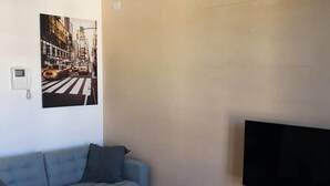 A room with a grey sofa, a flat-screen TV mounted on the wall, and a painting of a city street. - Two-room apartment overlooking the Basilica (Loreto)
