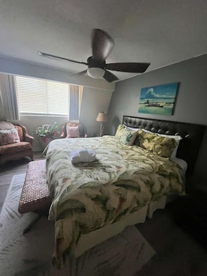 Une chambre à coucher avec un lit, un ventilateur de plafond, une fenêtre avec des stores, une chaise et un tableau accroché au mur.
