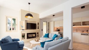 Ein modernes Wohnzimmer mit Sofa, Sesseln und Fernseher, angrenzend an eine Küche mit Schränken und Geschirrspüler.