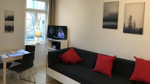 Olohuoneessa on musta sohva, punaiset koristetyynyt, jalustalla oleva televisio, työpöytä tuolilla sekä ikkuna, jossa on verhot.